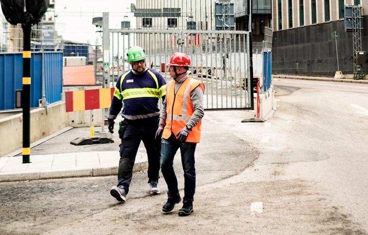 Två byggarbetare i skyddskläder och hjälmar går och samtalar på en gata intill en byggarbetsplats med metallstängsel och avspärrningar i bakgrunden.